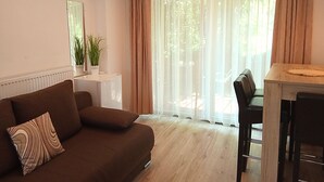 A living room with a brown sofa, a small white side table with two potted plants, a dining table with chairs, and a large window with sheer curtains. - Aparthotel Orient (Eger)
