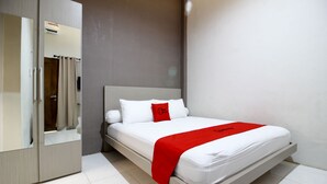 Un lit bien fait, avec du linge de lit blanc et un coussin rouge, une armoire en bois et un miroir.