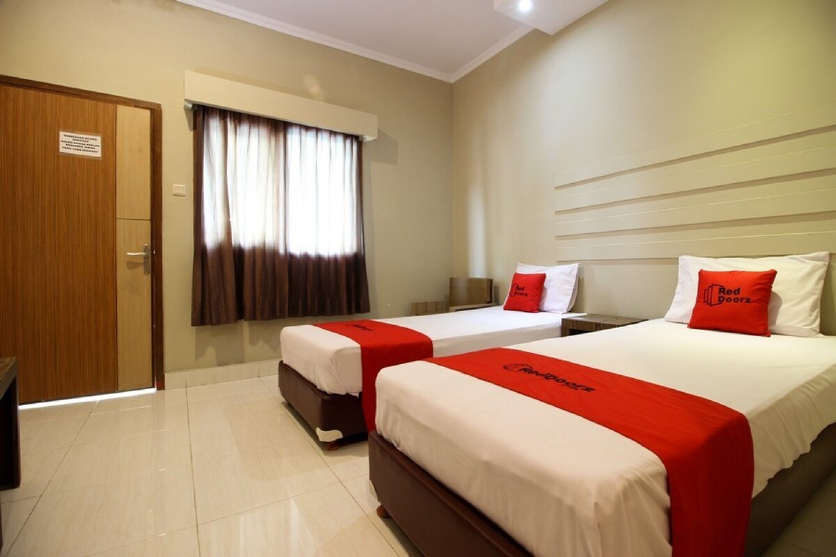 Une chambre d’hôtel avec deux lits, chacun recouvert d’une couverture rouge et blanche, une porte en bois et une fenêtre avec des rideaux.