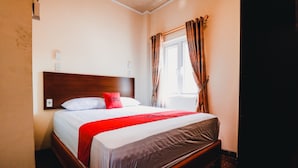 Un lit bien fait, avec une tête de lit en bois, un plaid rouge et une fenêtre avec des rideaux.