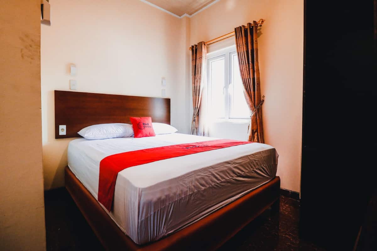 Un lit bien fait, avec une tête de lit en bois, un plaid rouge et une fenêtre avec des rideaux.