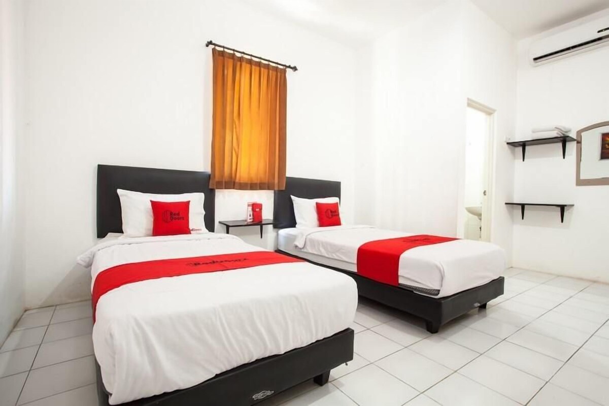 A hotel room with two beds, each with white bedding and red accents, a window with orange curtains, and a white tiled floor.