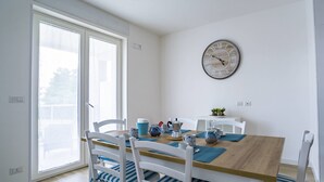 A dining area with a wooden table, white chairs with blue cushions, a wall clock, and a window with blinds. - La Casa del Cardinale (Monopoli)