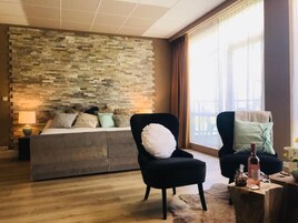 Una habitación de hotel moderna con pared de piedra, una cama, dos sillones, una pequeña mesa de madera y un gran ventanal con cortinas.
