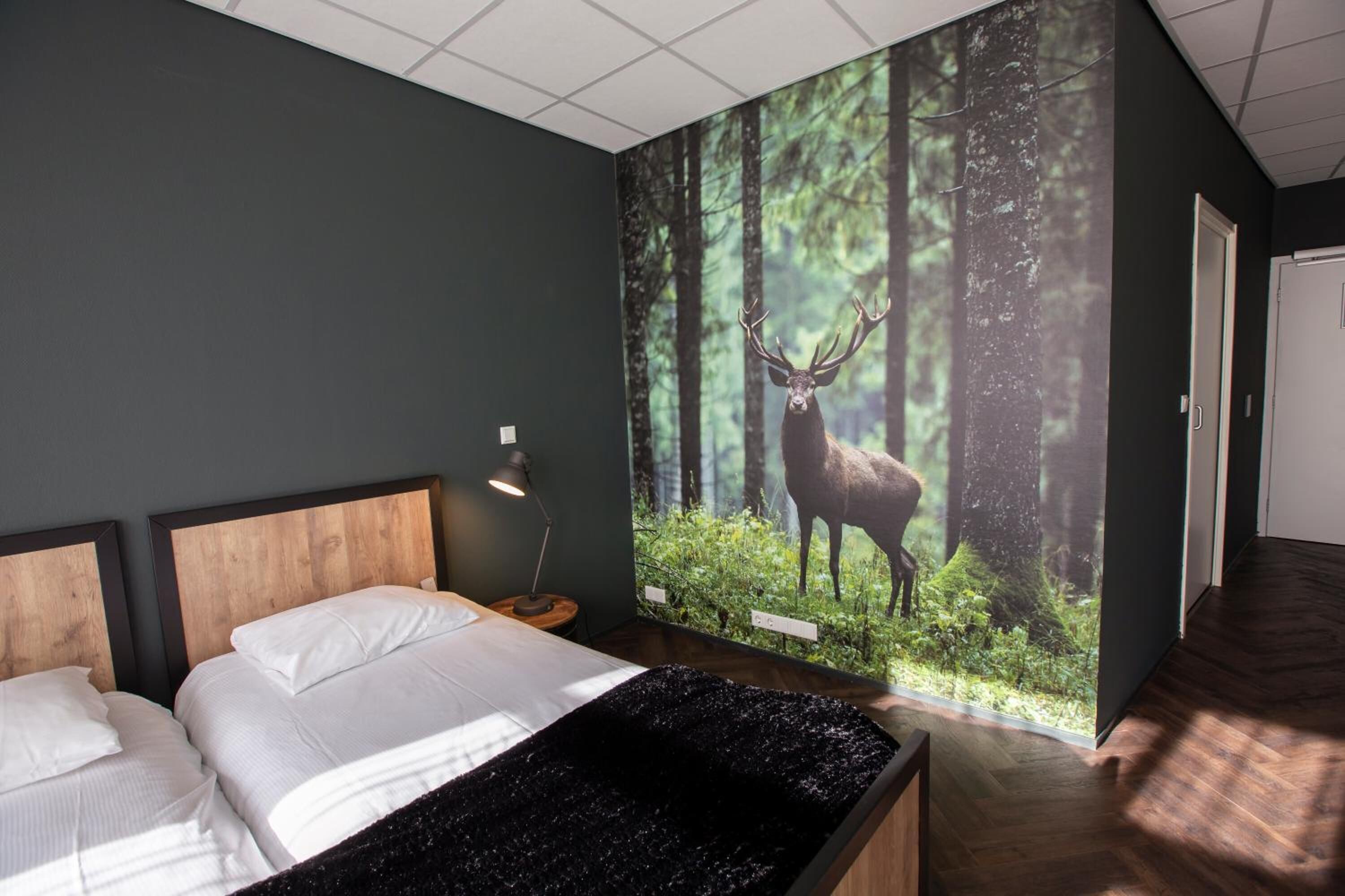 Un dormitorio con cama, mesita de noche, lámpara y un gran mural en la pared con un ciervo en un bosque.