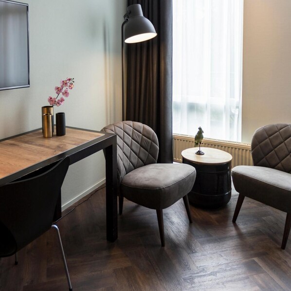 Una habitación moderna con una mesa de comedor de madera, dos sillas tapizadas, una mesita redonda y una lámpara de pie, además de un ventanal con cortinas.