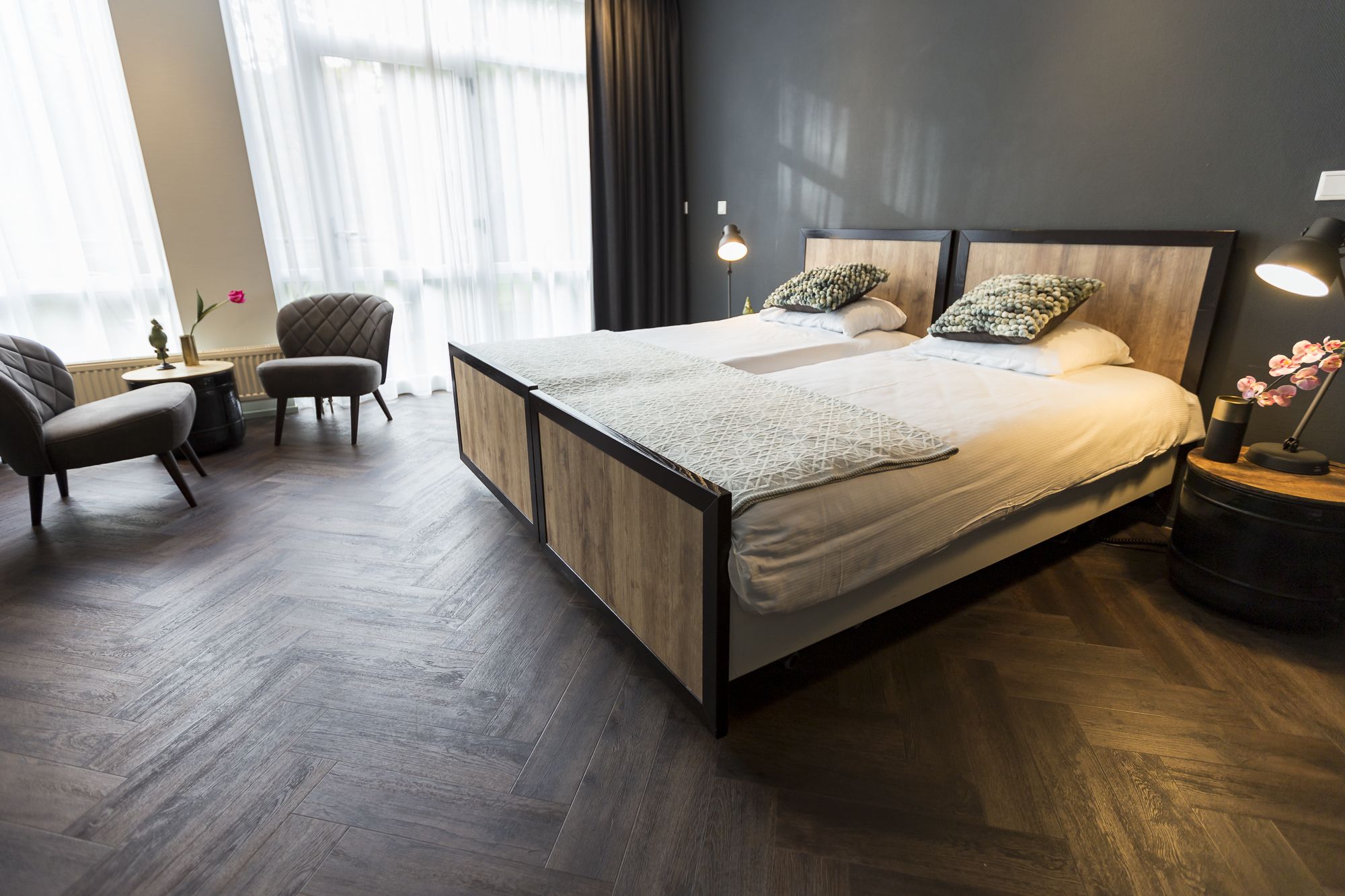 A modern hotel room with a large bed, two bedside tables, a chair, and a small table with a vase of flowers.
