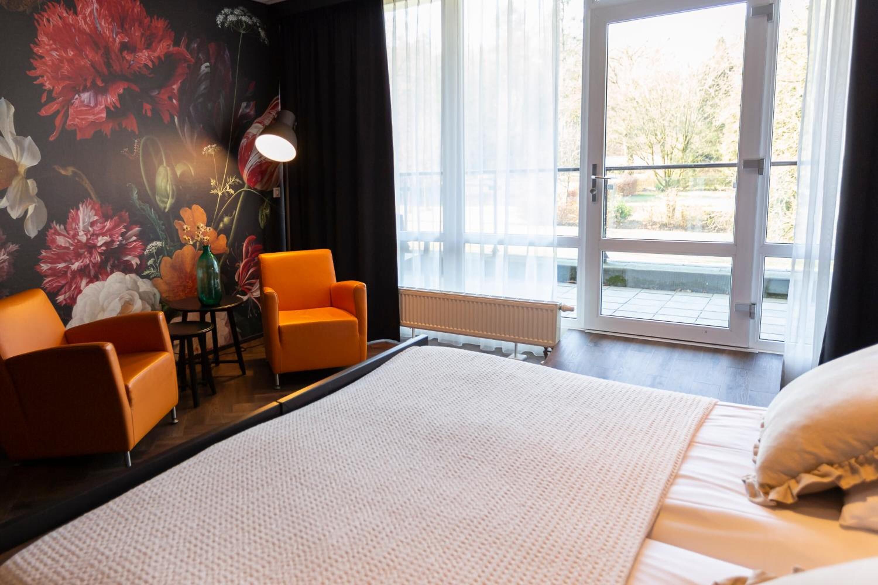 Habitación de hotel con una cama grande, dos sillones naranjas, una mesita con un florero y vistas al exterior a través de un ventanal.