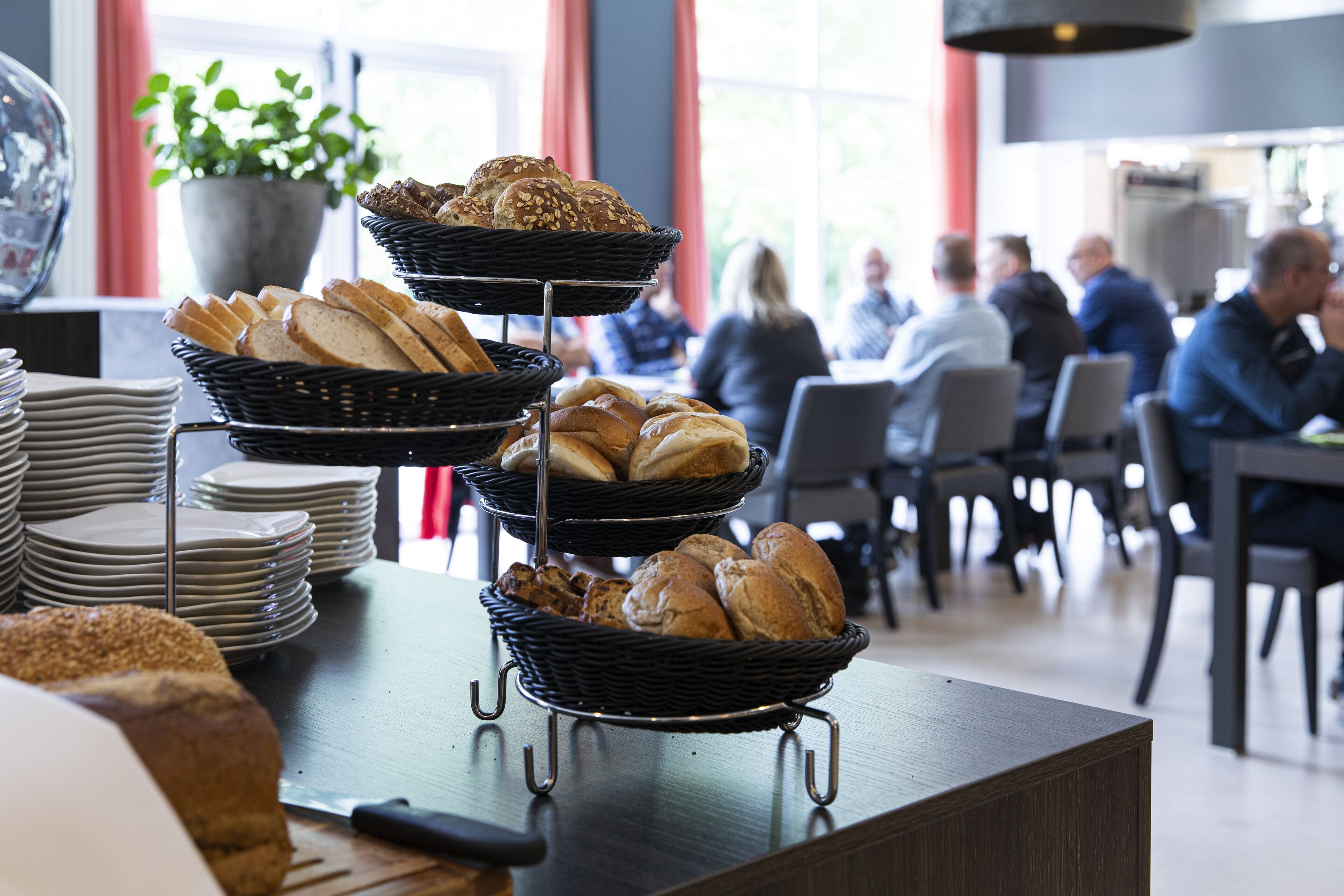 Ein reichhaltiges Buffet mit Brot und Gebäck in Körben, ein Messer auf einem Schneidebrett und eine Topfpflanze im Hintergrund.