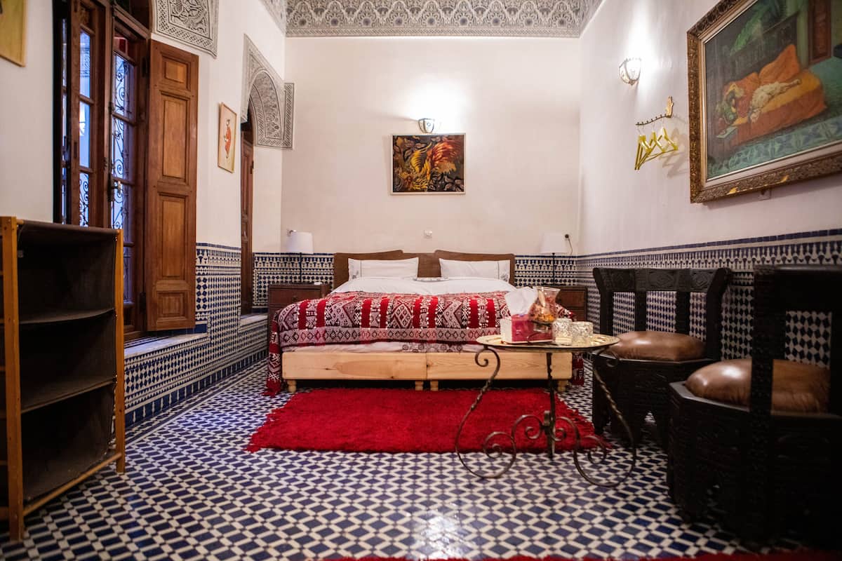 Quarto com piso estampado, cama com colcha vermelha e branca, cadeira de madeira e estante.