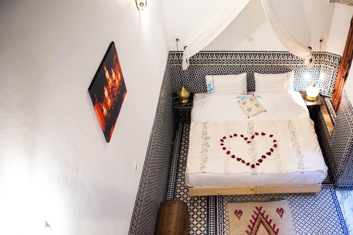 Uma cama com decoração em formato de coração, um criado-mudo com abajur e um piso estampado.