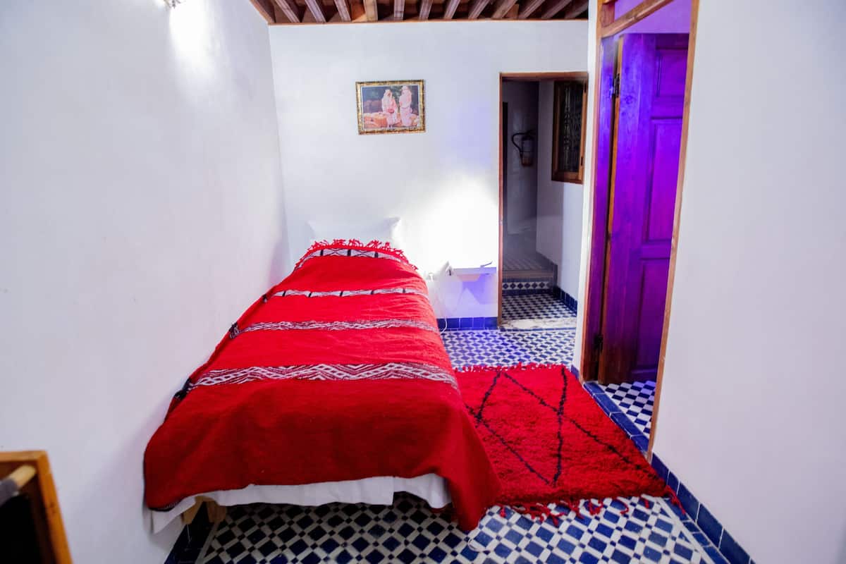 Um quarto pequeno com uma cama, um cobertor vermelho, chão quadriculado, porta com iluminação roxa e uma pequena moldura na parede.