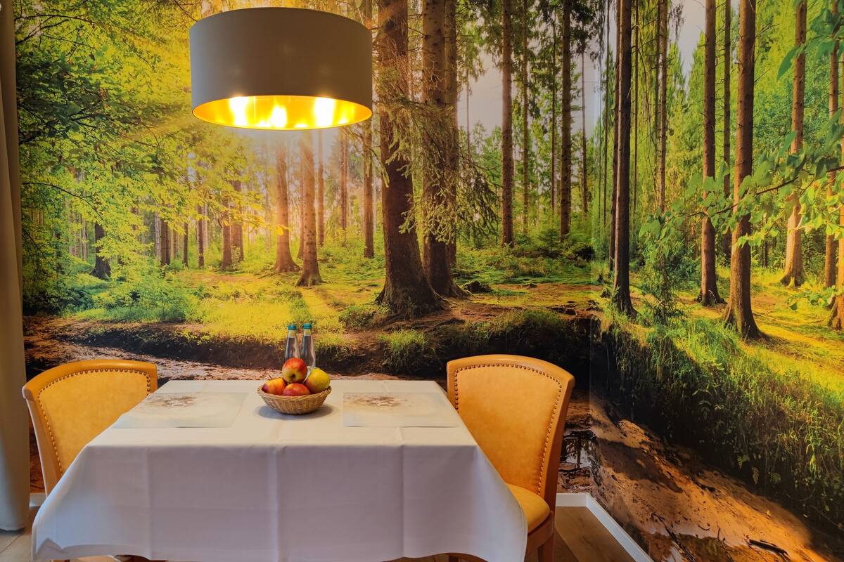 Un comedor con una mesa dispuesta para dos personas, un mural de un bosque en la pared y una lámpara colgante.