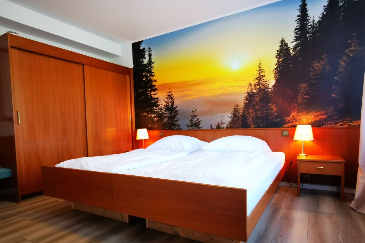 Un dormitorio con una cama de madera, dos mesitas de noche con lámparas, un armario y un mural que representa un bosque al atardecer.