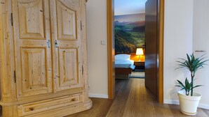 Ein Holzkleiderschrank mit mehreren Türen, ein Flur, der in ein Schlafzimmer mit Bett, Nachttisch, Lampe und Topfpflanze führt.
