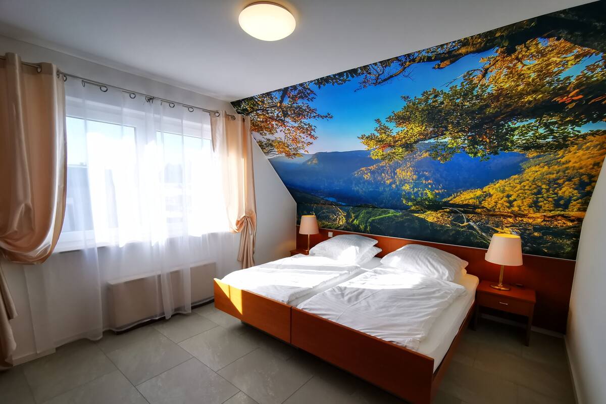 Un dormitorio con una cama grande, dos mesitas de noche con lámparas, una ventana con cortinas traslúcidas y un mural con una escena de bosque en la pared.