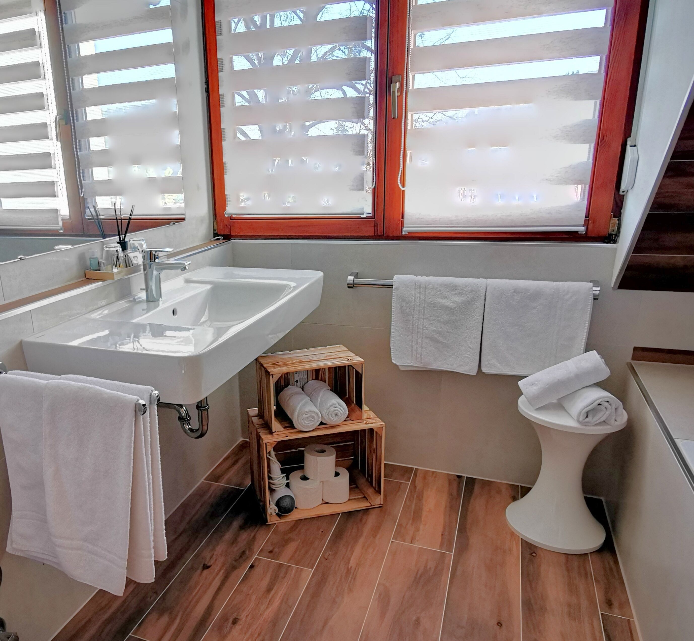 A bathroom with a white sink, wooden vanity, and a toilet. There are towels hanging on the wall and a wooden box with towels and toilet paper.