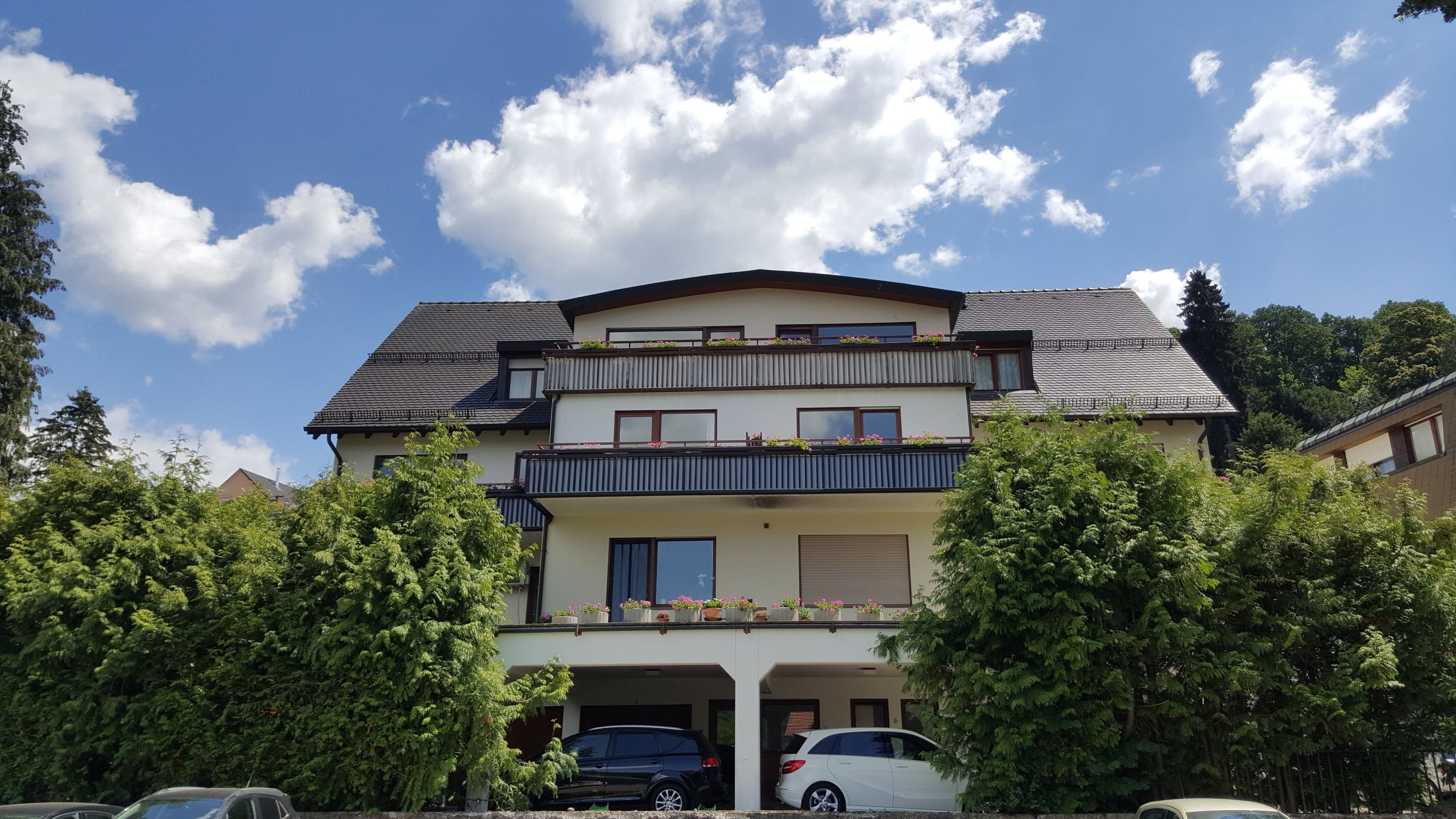 A two-story residential building with a balcony on the upper floor, a garage with two cars, and surrounded by trees.