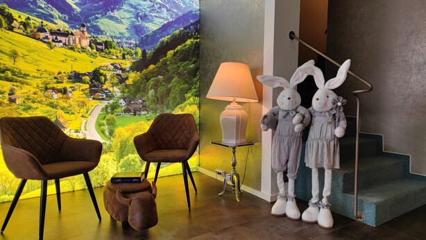 Ein Zimmer mit einer großen Wandmalerei, zwei braunen Sesseln, einer Lampe, einem kleinen Tisch, einem Stofftier und einer Treppe mit zwei weißen Hasenfiguren.