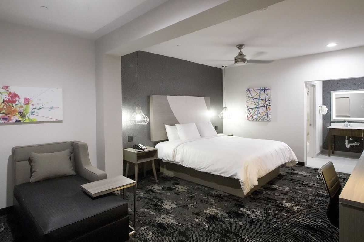 Una camera d'albergo moderna con un letto grande, un divano, una scrivania e un bagno con lavandino e specchio.