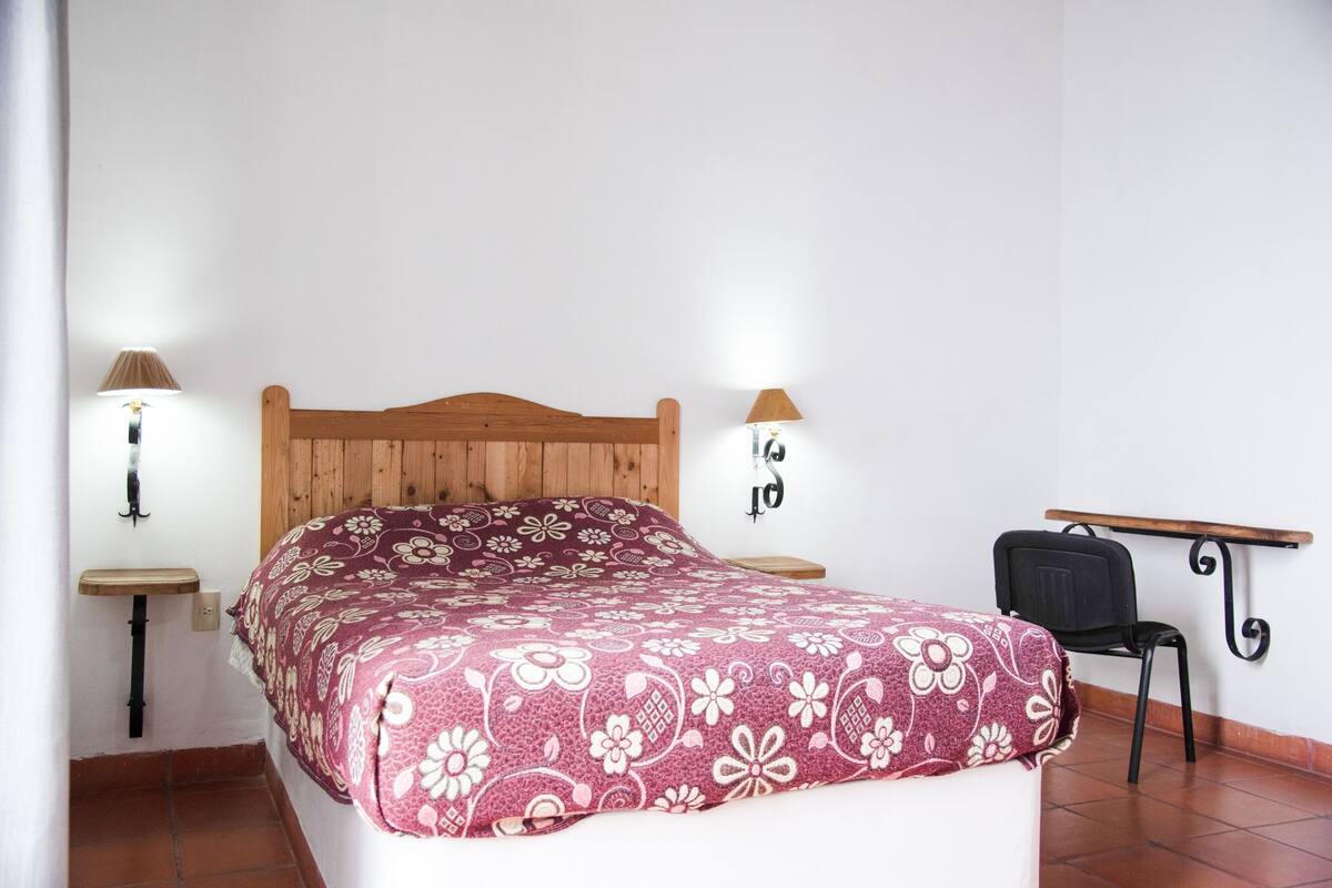 Une chambre à coucher avec un lit en bois, deux tables de chevet avec des lampes, une chaise et une petite étagère.
