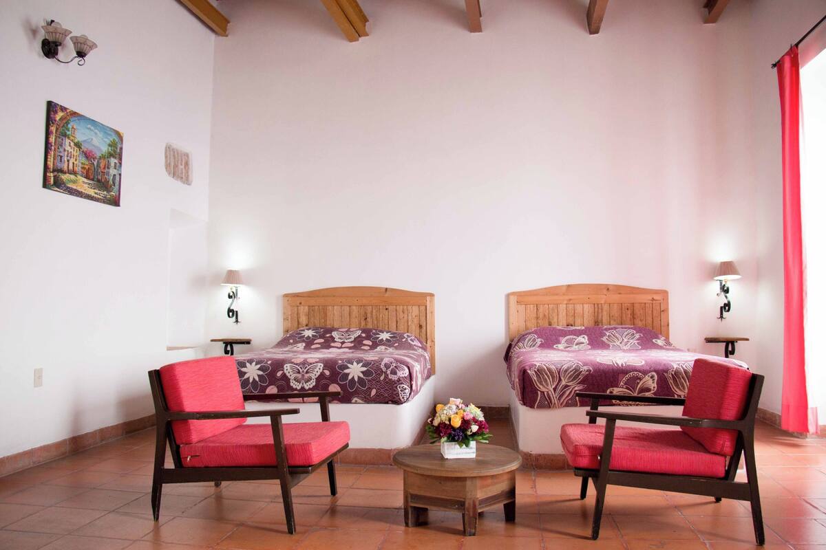 Une chambre avec deux lits, une petite table sur laquelle est posé un vase de fleurs, et deux chaises.