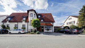 A two-story building with a red-tiled roof, a small garden with plants, and a car parked in front.
