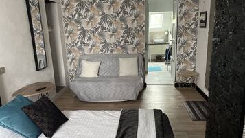 A modern bedroom with a grey sofa, a bed with a white and black bedspread, and a tropical leaf-patterned wallpaper.