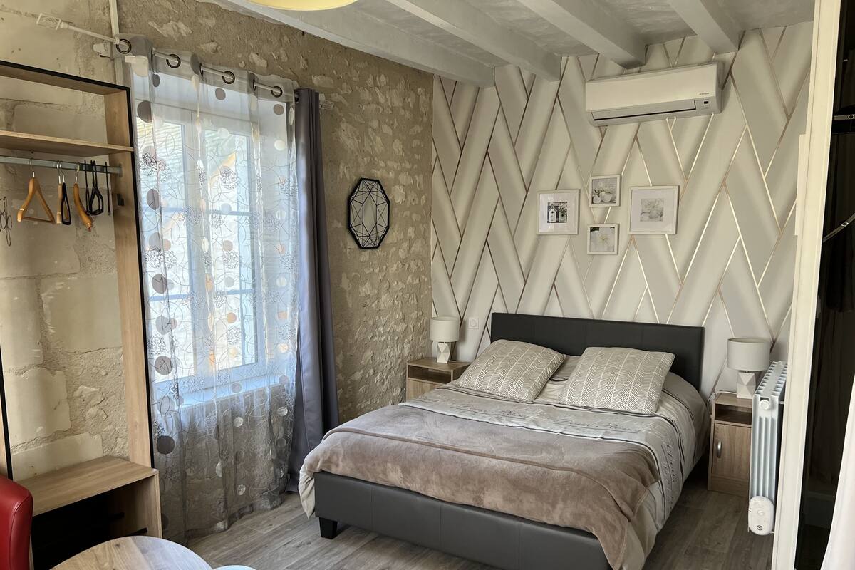 A bedroom with a bed, a nightstand, a lamp, a radiator, a window with curtains, and a wall with a geometric pattern.