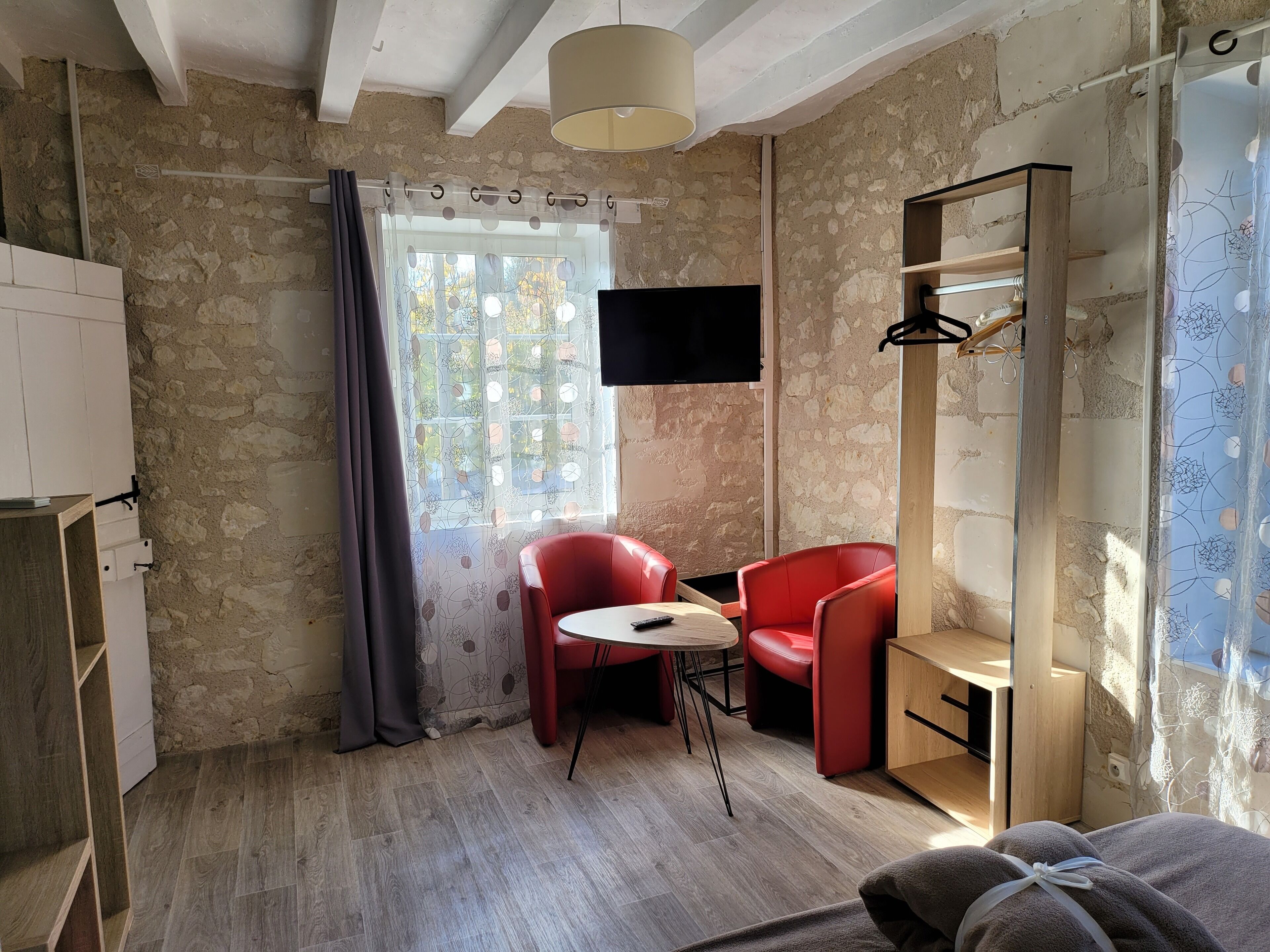 Une pièce avec un mur en pierre, un plancher en bois, une chaise rouge, une petite table, un téléviseur fixé au mur et une fenêtre avec un rideau.