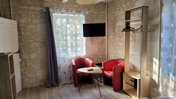 Une pièce avec un mur en pierre, un plancher en bois, une chaise rouge, une petite table, un téléviseur fixé au mur et une fenêtre avec un rideau.