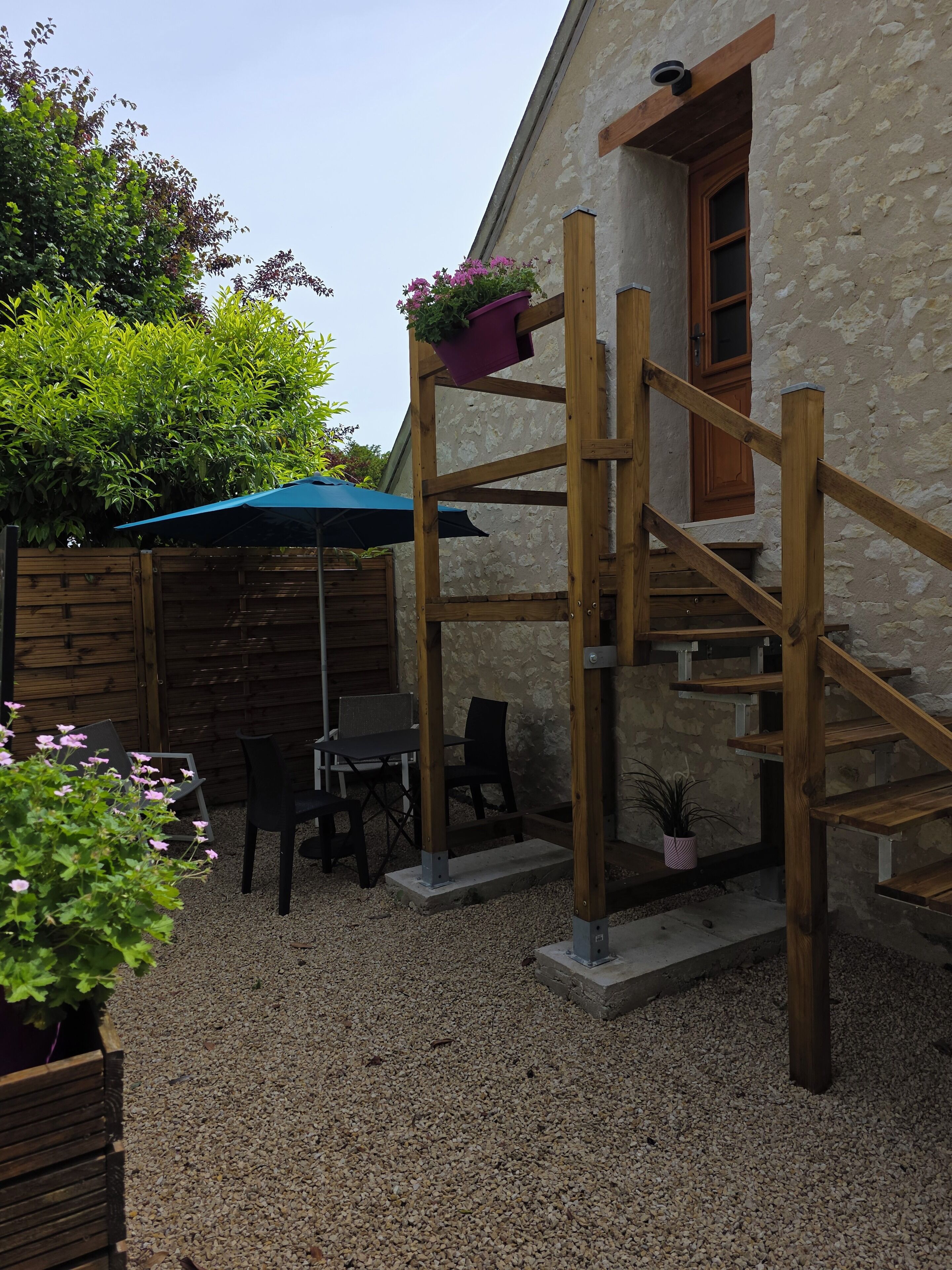 Un escalier extérieur en bois mène à un bâtiment en pierre, qui est entouré de gravier. On y trouve une table et des chaises sous un parasol bleu.