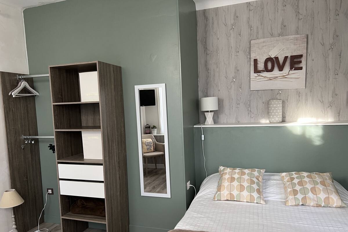 A bedroom with a bed, a wardrobe, a mirror, a wall-mounted shelf with a lamp, and a 'LOVE' sign.