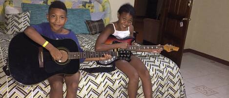 Two individuals seated on a bed with patterned bedding, each holding a guitar.