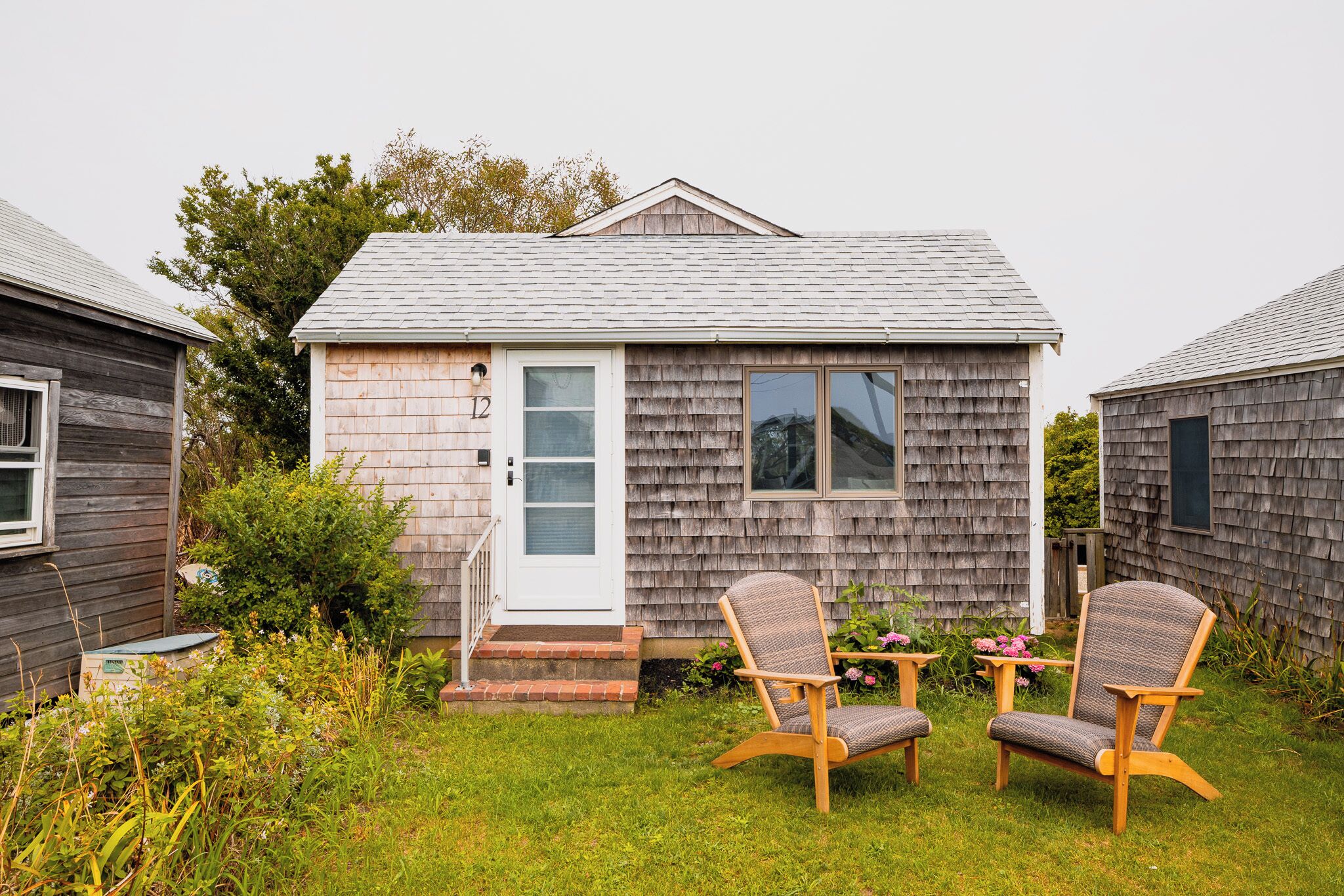 A small wooden house with a door and windows, a grassy yard, and two wicker chairs.