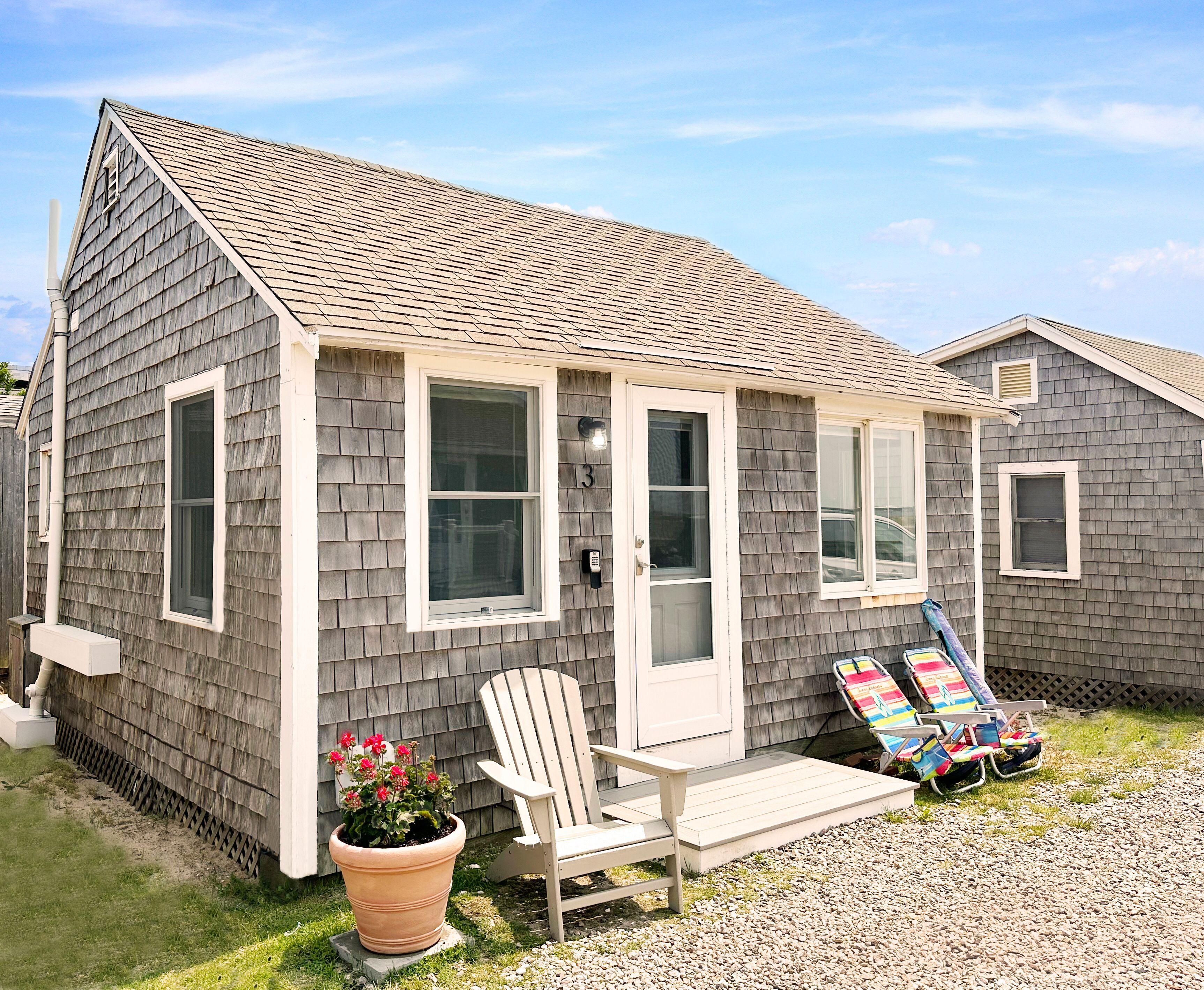 A single-story house with a gray shingle roof, white trim, and a front porch. There are two windows on the front, a door, and a small porch area with a chair and a potted plant.