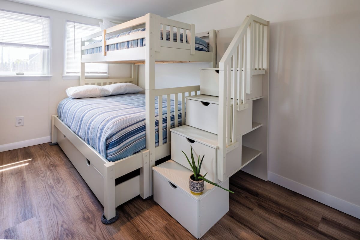 A bunk bed with a built-in desk and shelving, a potted plant, and a window with blinds.