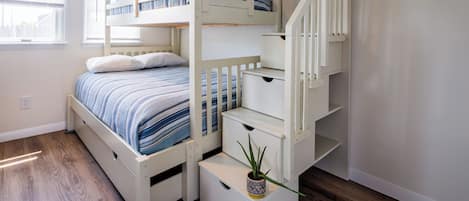 A bunk bed with a built-in desk and shelving, a potted plant, and a window with blinds.