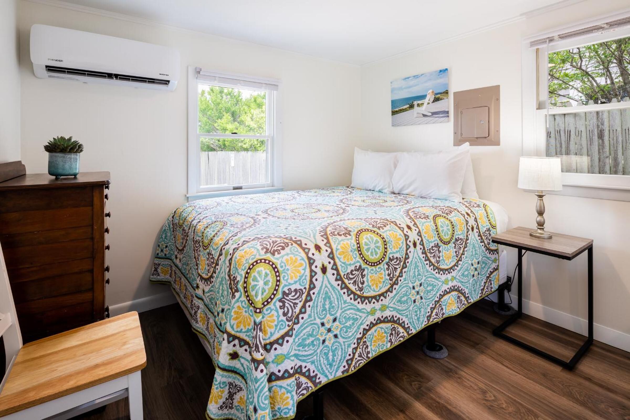 A bedroom with a bed, a bedside table with a lamp, a dresser, a window with a view, and an air conditioner.
