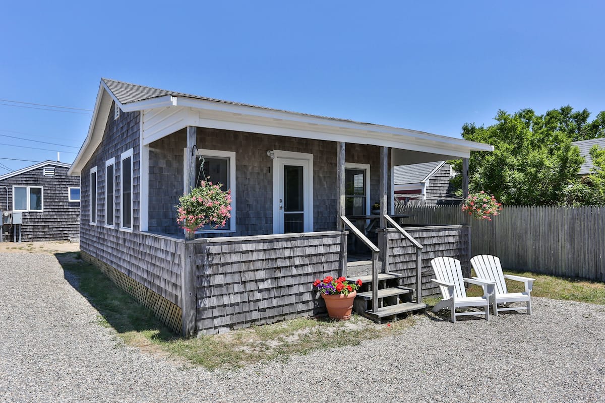 A small, single-story building with a porch, two potted plants, and two Adirondack chairs.