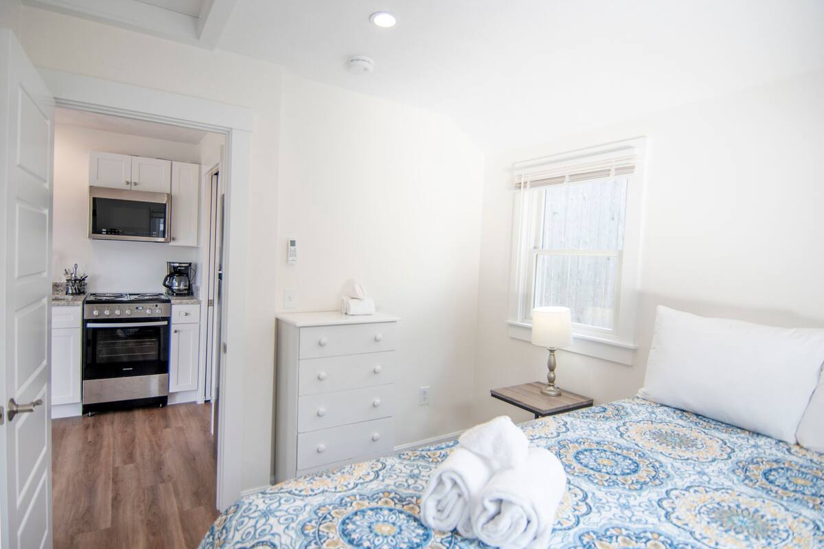 A bedroom with a bed, a bedside table, a lamp, a window, and a view of a kitchen with a stove and microwave.
