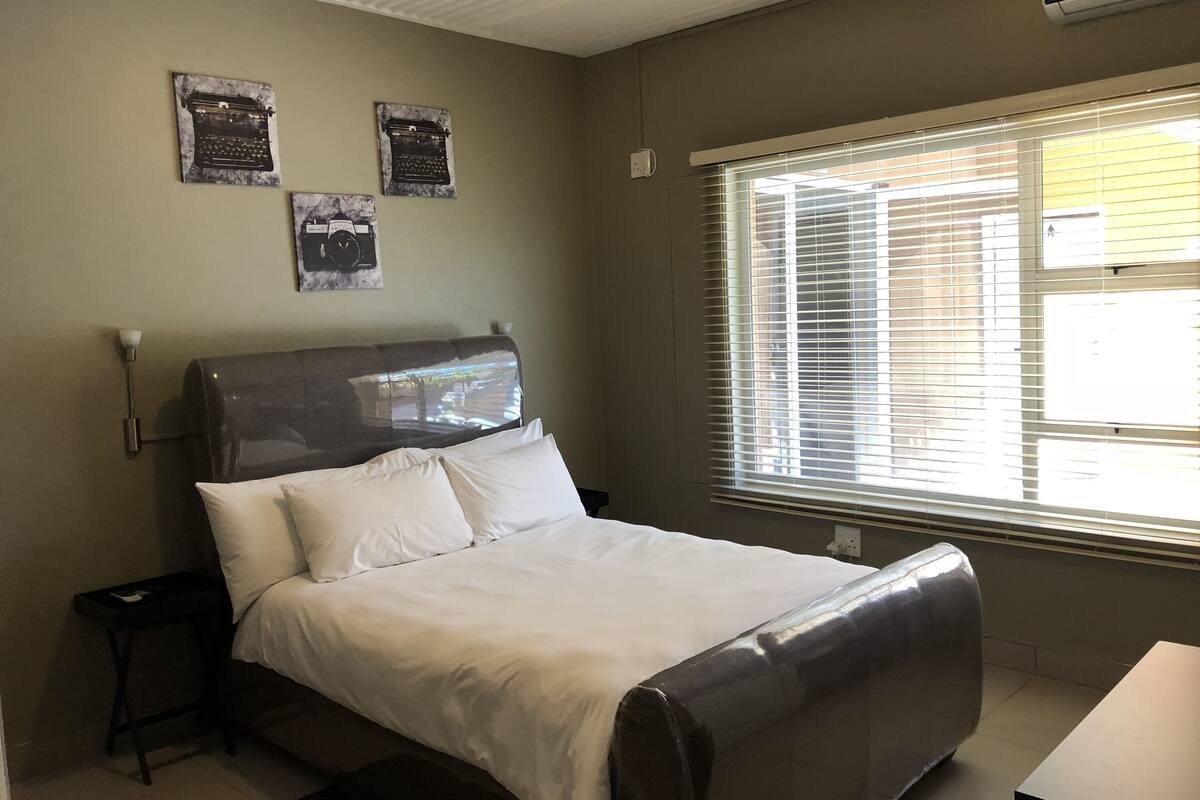 A bedroom with a bed, a nightstand, a window with blinds, and wall-mounted artwork.