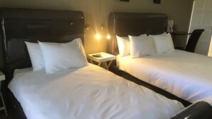 Habitación de hotel con dos camas, cada una con ropa de cama blanca y cabeceros. Hay una mesita de noche pequeña con una lámpara y un teléfono.