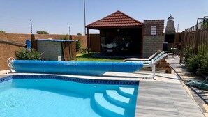 Una piscina con solárium de baldosas, un área de estar cubierta y una estructura de ladrillo al fondo.