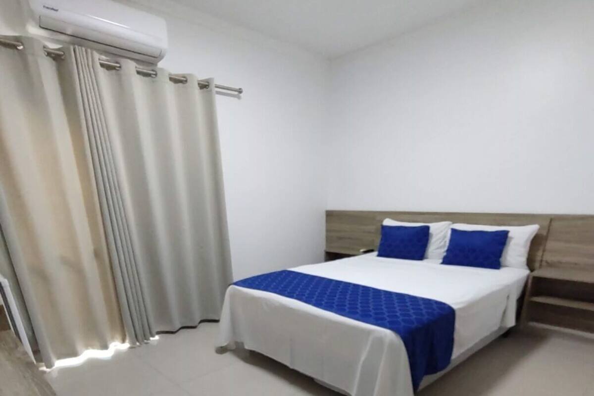 Una habitación de hotel con cama, cortinas, cabecero de madera y unidad de aire acondicionado blanca.
