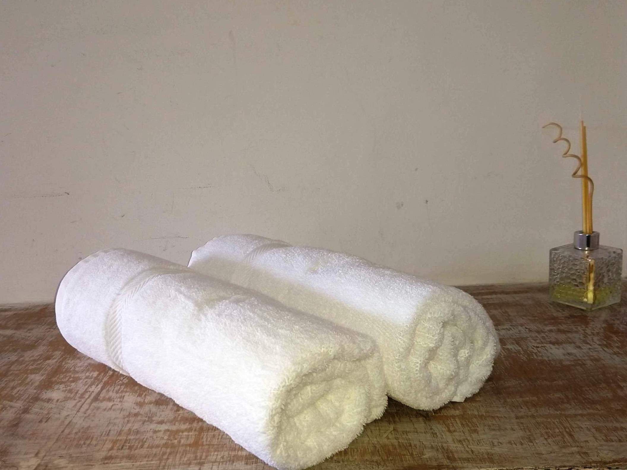 Dos toallas blancas enrolladas sobre una superficie de madera, con una botella de vidrio que contiene un líquido amarillo y un recipiente metálico con un objeto amarillo en una mesa, frente a una pared lisa.