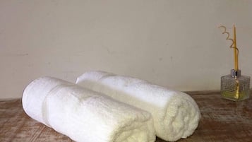 Deux serviettes blanches roulées sont posées sur une surface en bois, à côté d’une bouteille en verre contenant un liquide jaune et d’un récipient en métal contenant un objet jaune, le tout sur une table devant un mur uni.