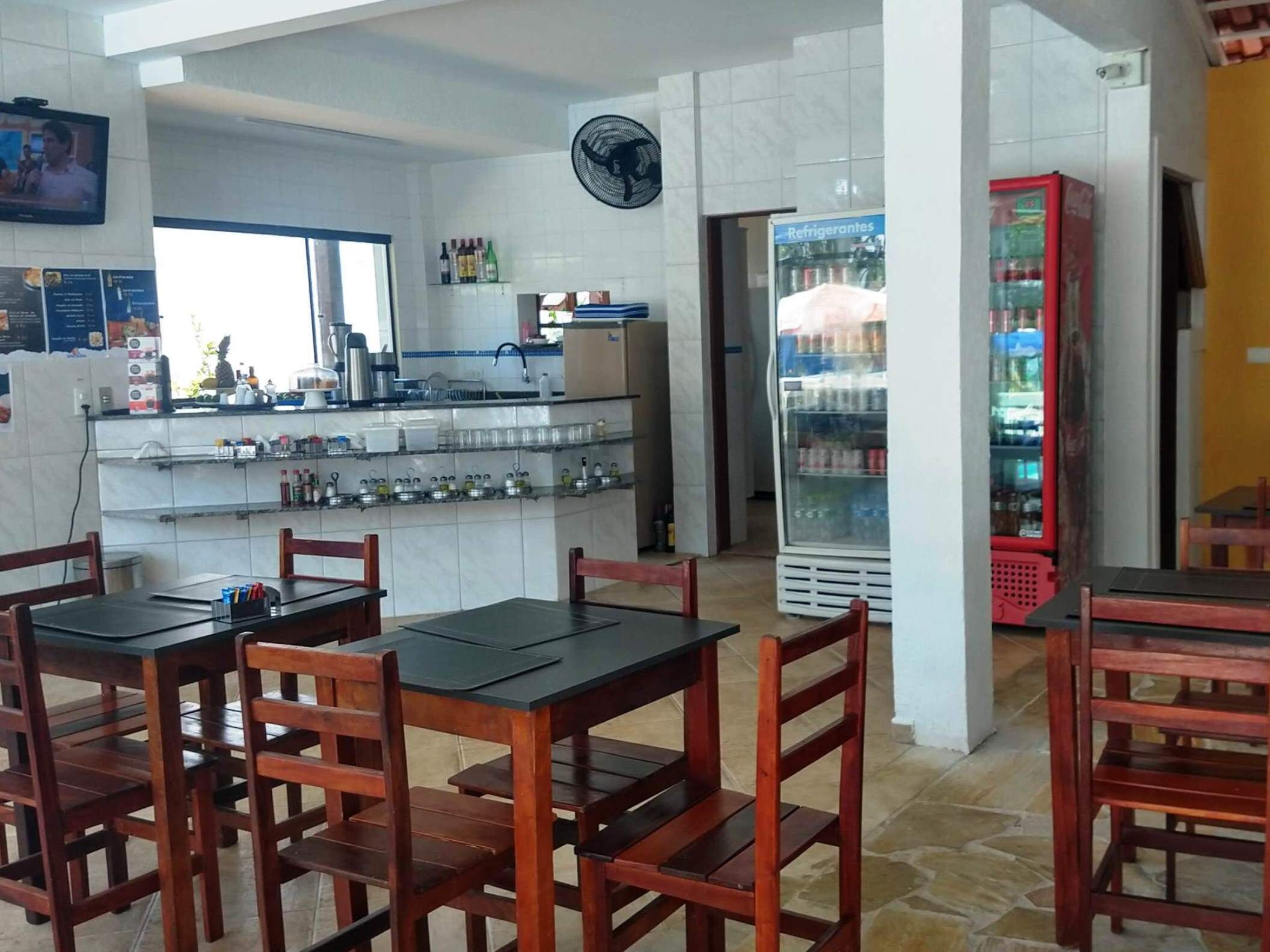 A restaurant interior with wooden tables and chairs, a kitchen area with a fan, and a refrigerator stocked with beverages.