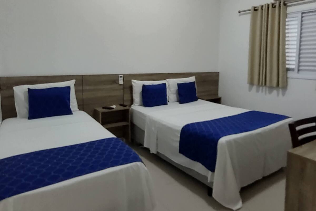 Una habitación con dos camas, cada una con cubrecama azul y ropa de cama blanca. Tiene un cabecero de madera, una mesita de noche con lámpara y una ventana con cortinas.