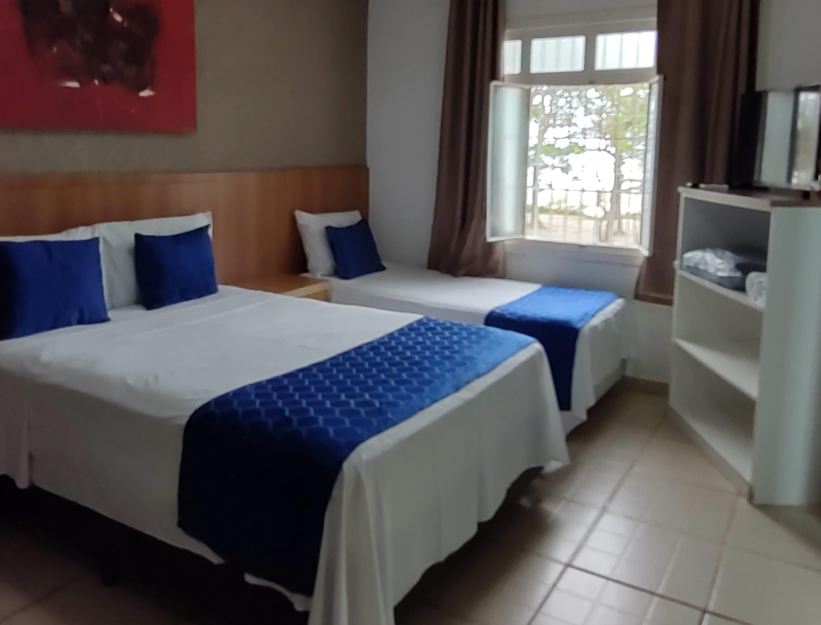Hotellihuone, jossa on kaksi sänkyä, yöpöytä, ikkuna, jossa on verhot, ja valkoinen hylly, jonka päällä on tavaroita.